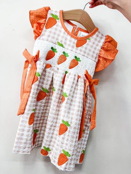 The Carrot Patch Twirl Dress by Tiny Tot Bundles features orange ruffled sleeves, side bows, and a white and peach checkered pattern with orange carrot designs, displayed on a wooden hanger.