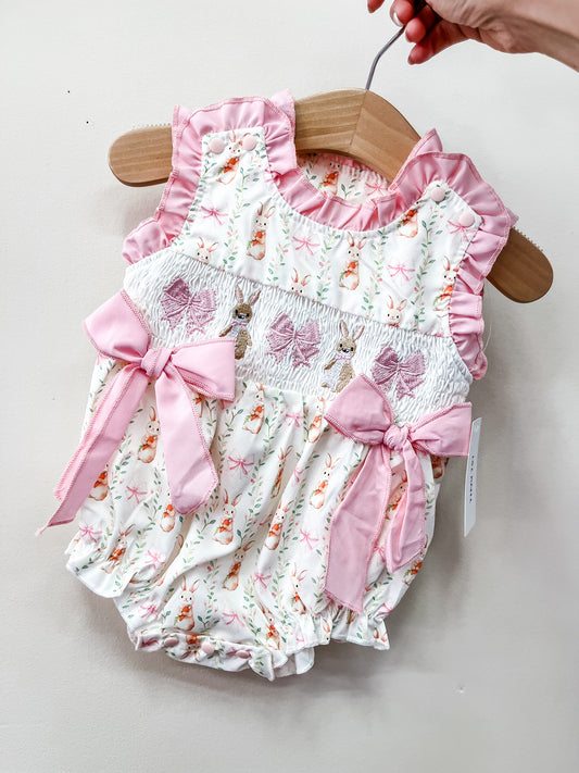 The Blushing Bunny Bow Bubble by Tiny Tot Bundles features a pink and white bunny pattern, pink ruffled trim, and two pink bows. Displayed on a wooden hanger held by hand against a plain background.