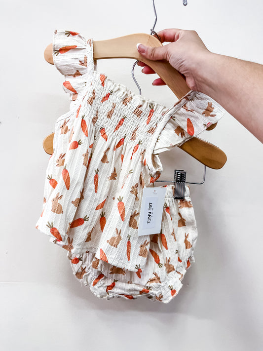 A hand holds Tiny Tot Bundles’ Bunny Garden Patch Set—a white baby outfit with orange carrot and brown bunny print, featuring ruffled straps and a matching bloomer.