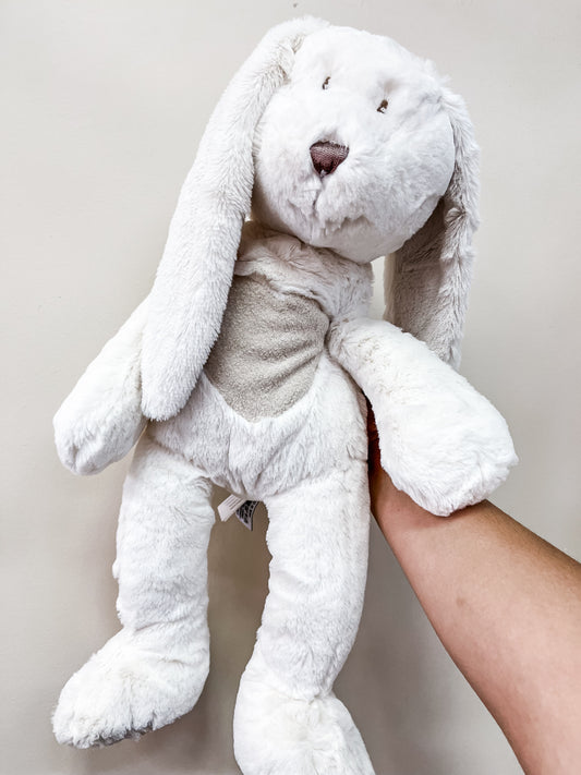 A person holds up the Tiny Tot Bundles White Bunny Heart Plush, featuring floppy ears and a gray heart-shaped patch on its belly, against a plain, light-colored background.