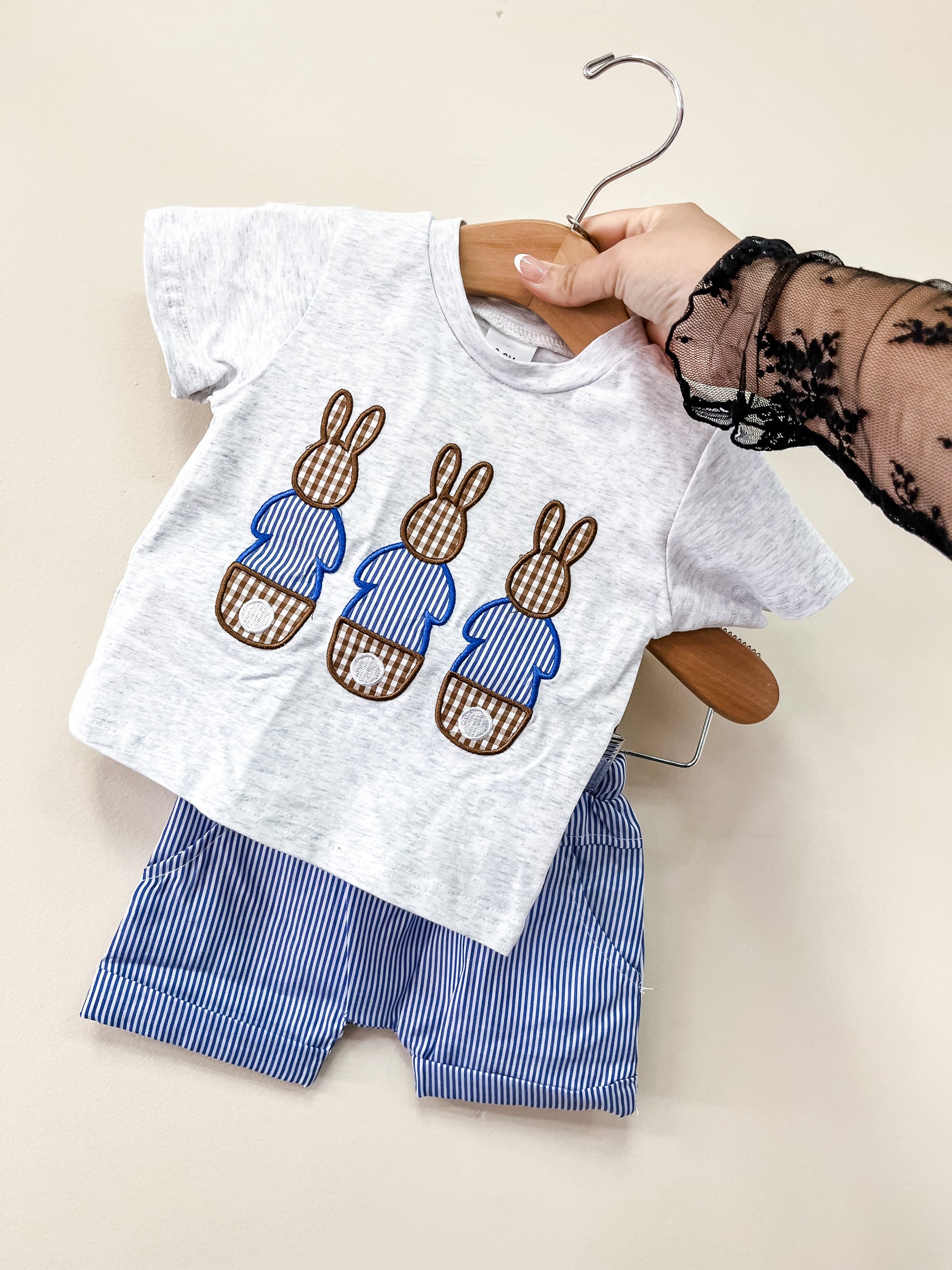 A hand holds up the Tiny Tot Bundles Gingham Bunny Boys Set, featuring a light gray t-shirt with three blue bunny designs and matching blue and white striped shorts.