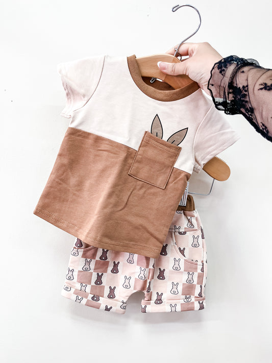 A hand displays the Tiny Tot Bundles Brown Checkered Boys Set: a short-sleeve shirt with a brown pocket and bunny ears, paired with matching shorts featuring a bunny face on a white and beige checkerboard pattern.