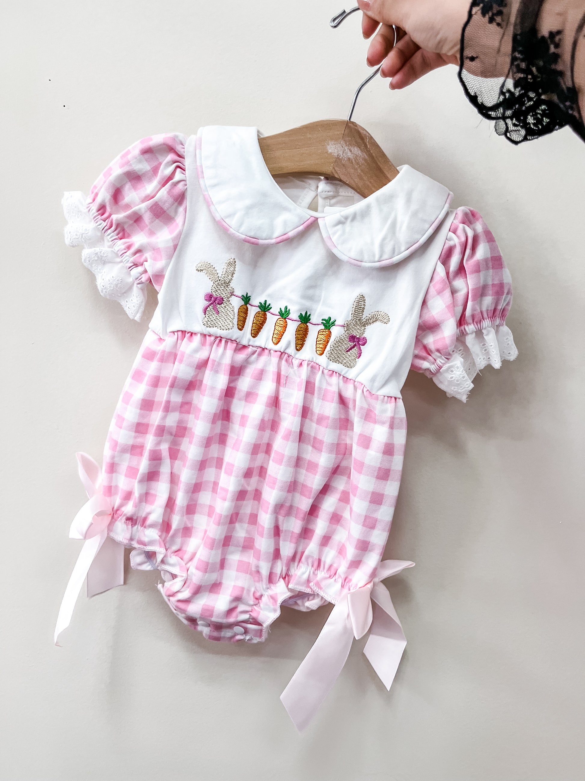 A hand holds Tiny Tot Bundles’ Bunny Patch Gingham Bubble, a pink and white check romper with puff sleeves, lace trim, a large collar, embroidered bunnies and carrots, plus pink ribbon bows on the legs.