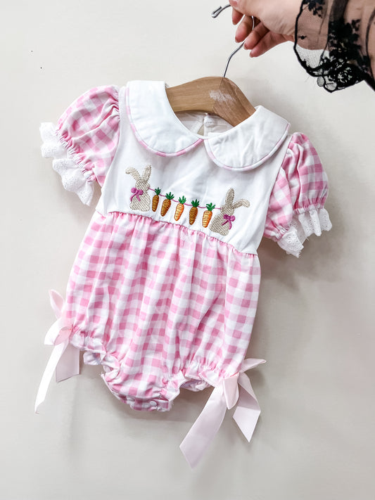 A hand holds Tiny Tot Bundles’ Bunny Patch Gingham Bubble, a pink and white check romper with puff sleeves, lace trim, a large collar, embroidered bunnies and carrots, plus pink ribbon bows on the legs.