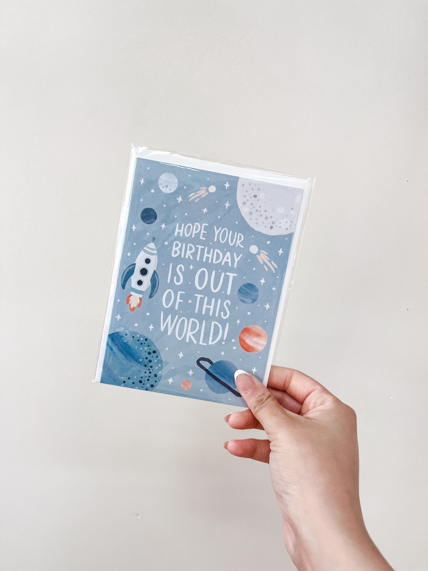 A hand holds the "Out of This World - Birthday Card" by love Light Paper, showing planets, stars, a rocket, and the message "Hope your birthday is out of this world!" on a light blue background.