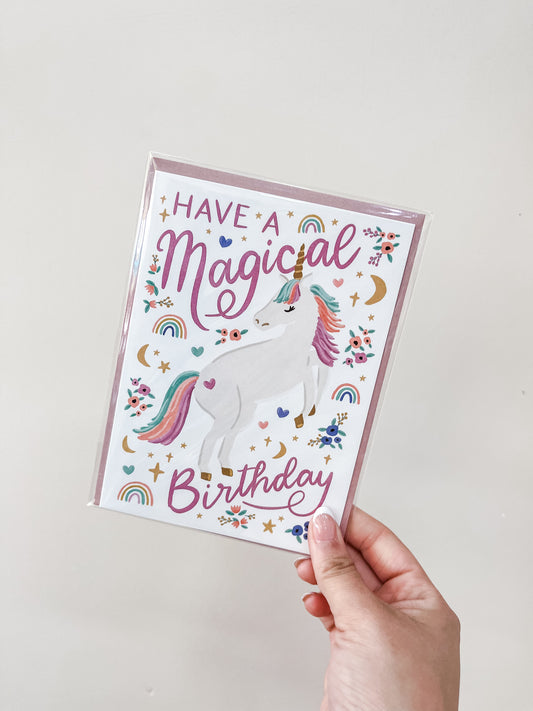 A hand holds the "Unicorn - Birthday Card" by love Light Paper, featuring a white unicorn with a rainbow mane and tail among stars, rainbows, hearts, and flowers. The card says "Have a Magical Birthday" in purple and pink text.