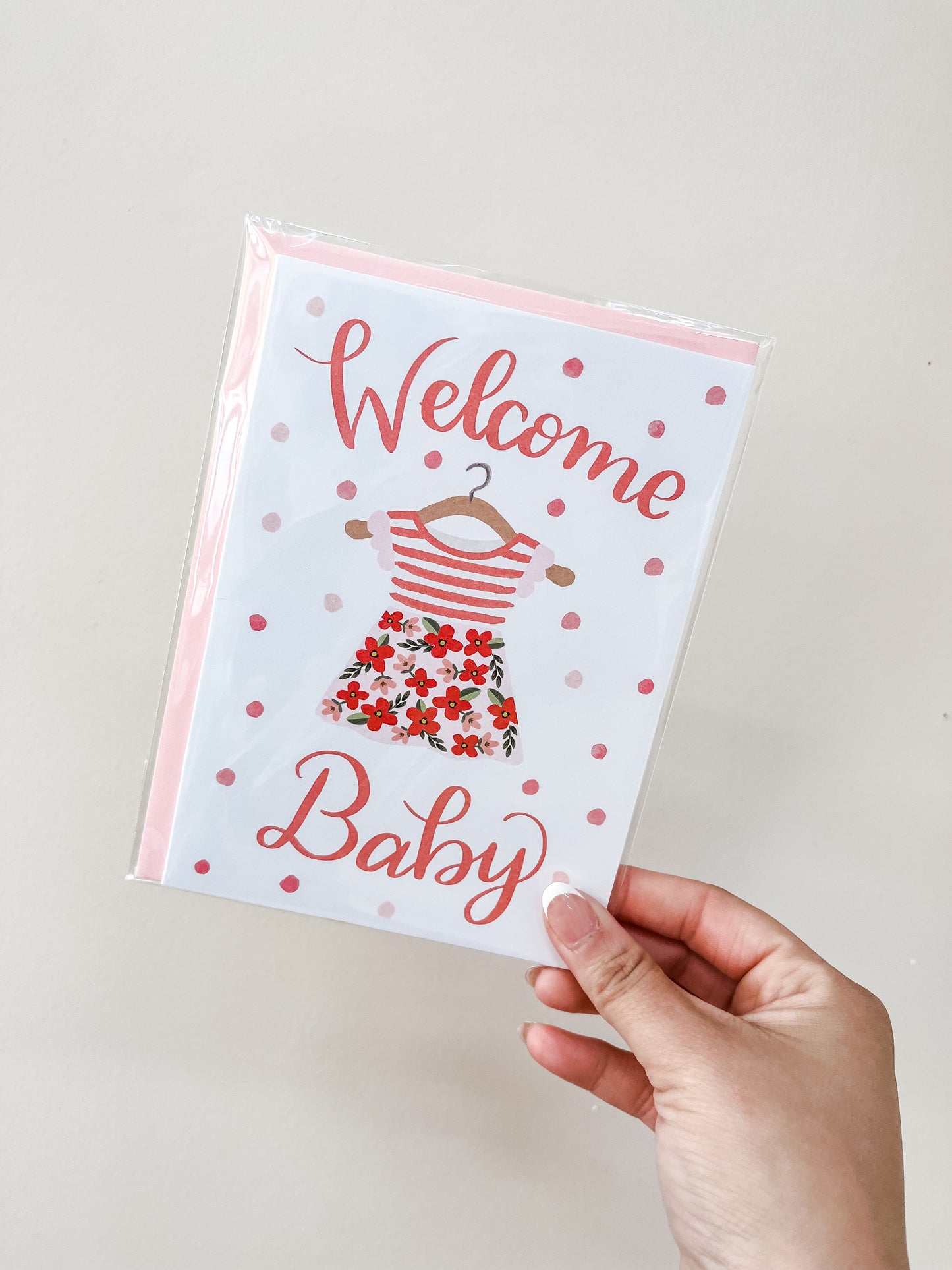 A hand holds the "Welcome Baby Girl - Card" by love Light Paper, featuring pink polka dots and a baby dress illustration with pink stripes and a floral skirt.