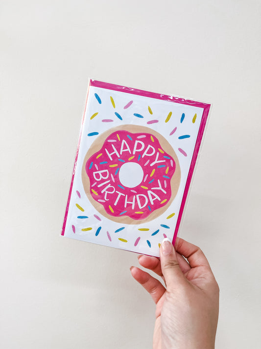 A hand holds the Donut - Birthday Card by love Light Paper, showing a pink frosted donut with colorful sprinkles and "HAPPY BIRTHDAY" across it on a plain white background.