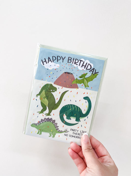A hand holds the love Light Paper Dinosaur - Birthday Card, featuring colorful cartoon dinosaurs, confetti, and a volcano with the message "Party like there's no tomorrow.