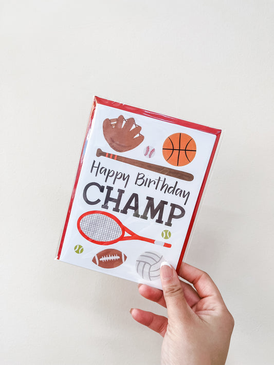 A hand holds the "Champ - Birthday Card" by love Light Paper, featuring sports gear like a baseball glove, basketball, football, tennis racquet and balls, and volleyball. The card displays the message "Happy Birthday CHAMP.