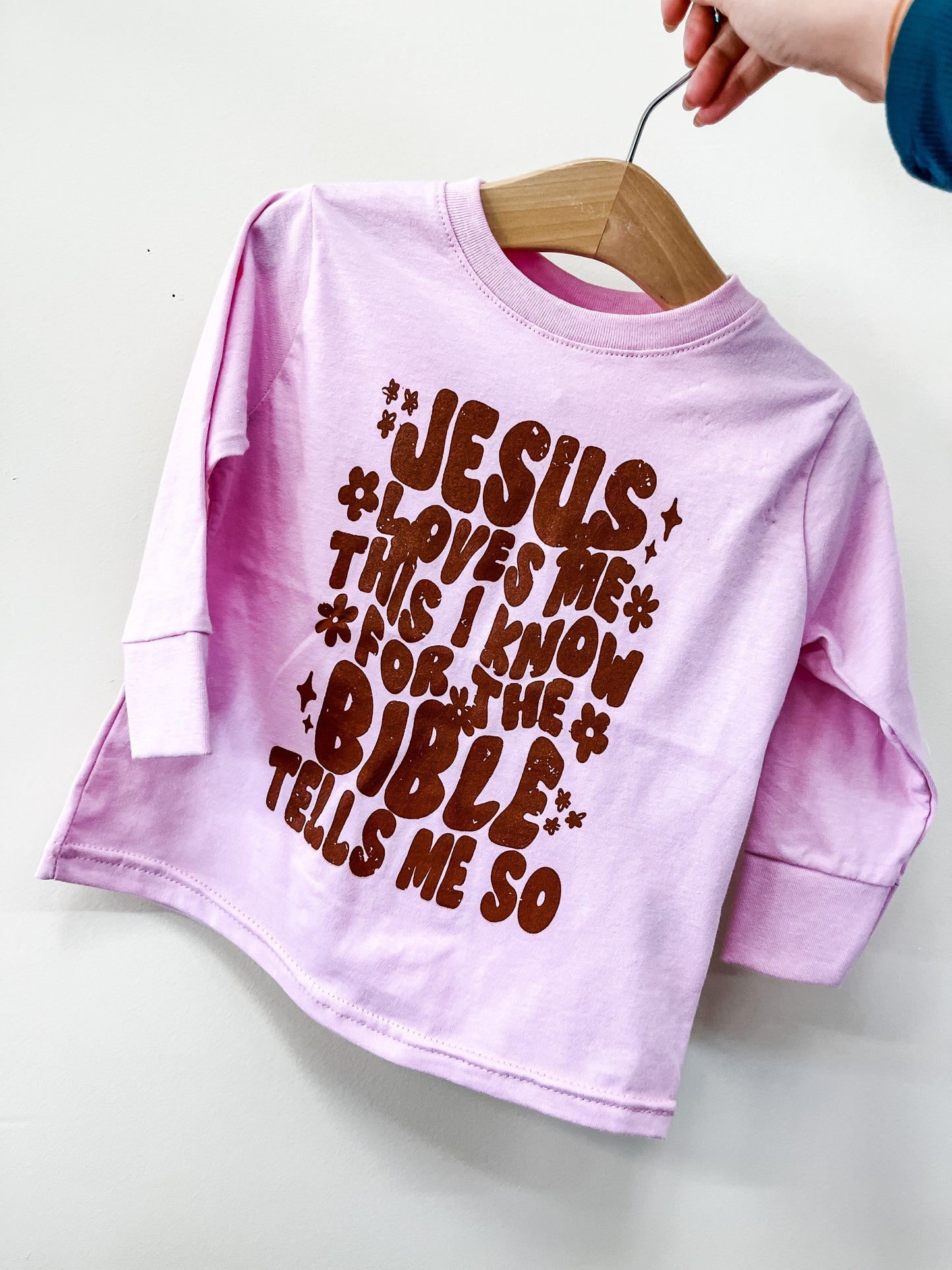 A Beauts & Beaus Jesus Loves Me Long Sleeve toddler shirt in light pink hangs on a hanger, featuring brown retro “Jesus loves me, this I know for the Bible tells me so” text. A hand holds the hanger.