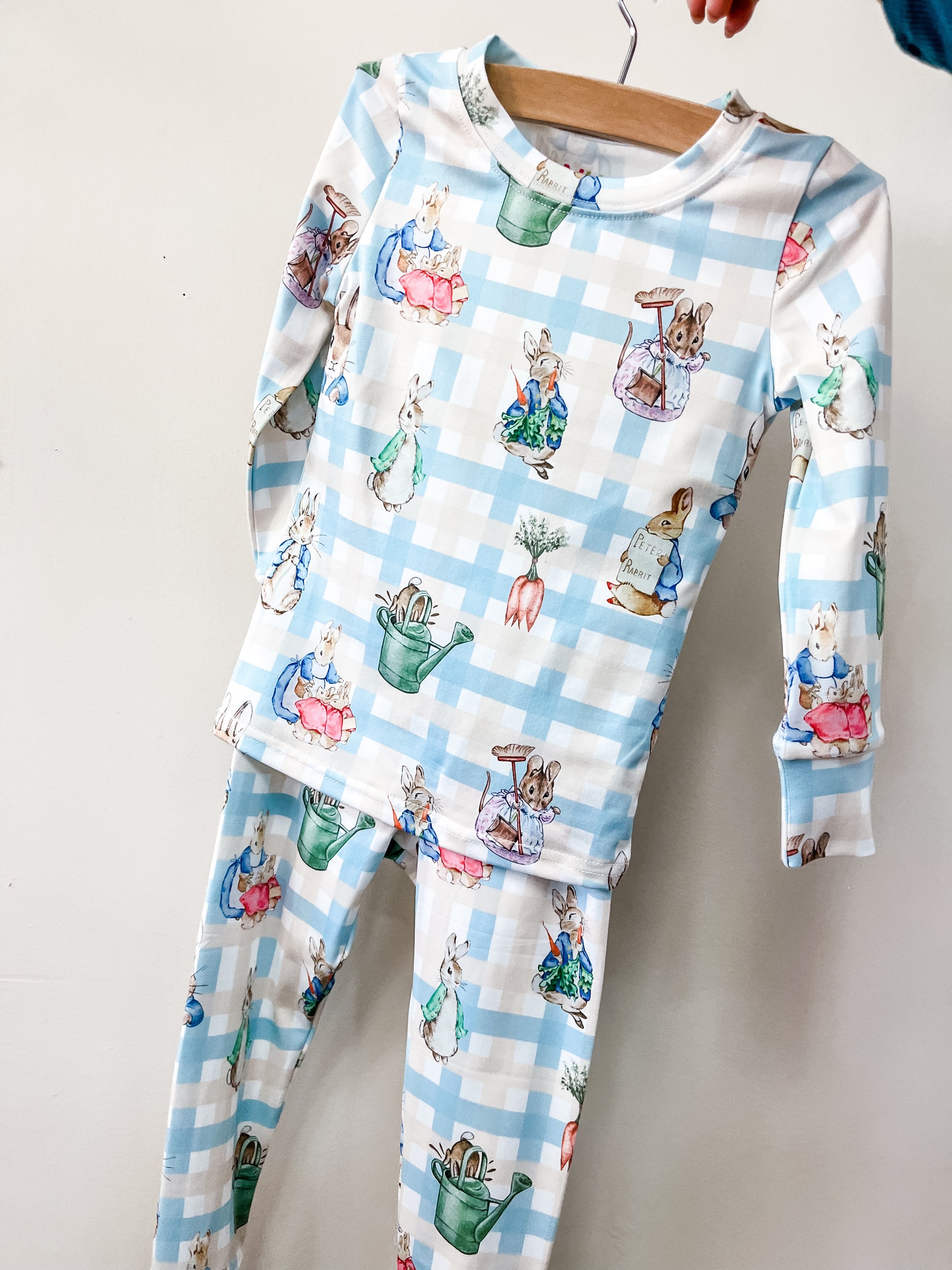 Tiny Tot Bundles' Peter's Easter Picnic Jammies Set features blue and white checkered children’s pajamas with rabbit and watering can prints, displayed on a wooden hanger against a plain background.