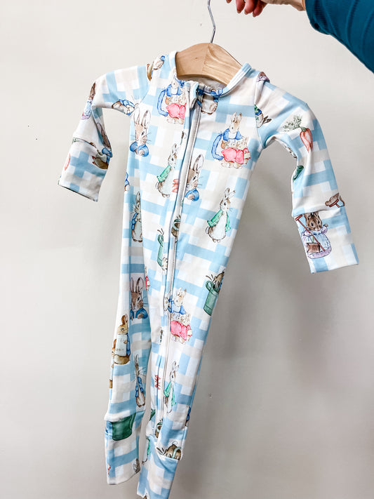 A hand holds up Tiny Tot Bundles' Peter’s Easter Picnic Long Zippy, a blue and white checkered baby onesie with long sleeves, front zipper, and adorable rabbit and woodland character illustrations.