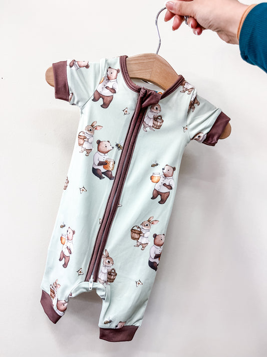 The Peter's Honey Adventure Shorty Zippy by Tiny Tot Bundles, a mint green baby romper with brown trim and cute bear and bunny prints, hangs on a wooden hanger held by a hand against a simple background.