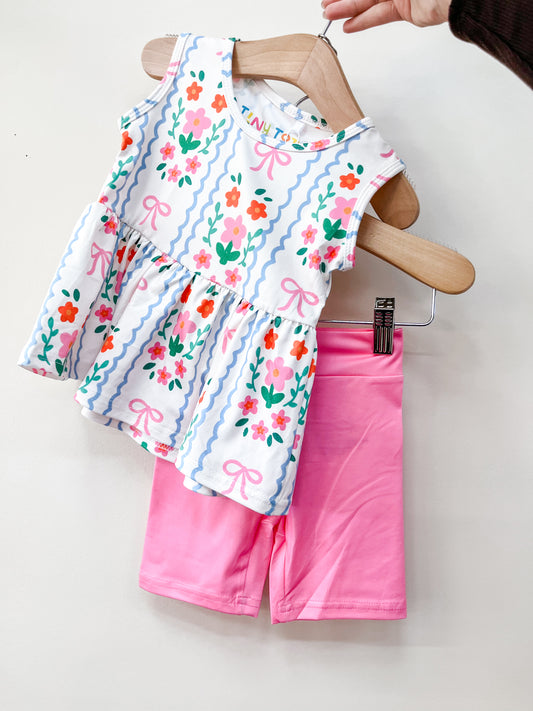 The Tiny Tot Bundles Ribbon Garden Peplum Set features a sleeveless white toddler dress with floral and bow designs on a wooden hanger, paired with bright pink shorts on another hanger, held against a white background.