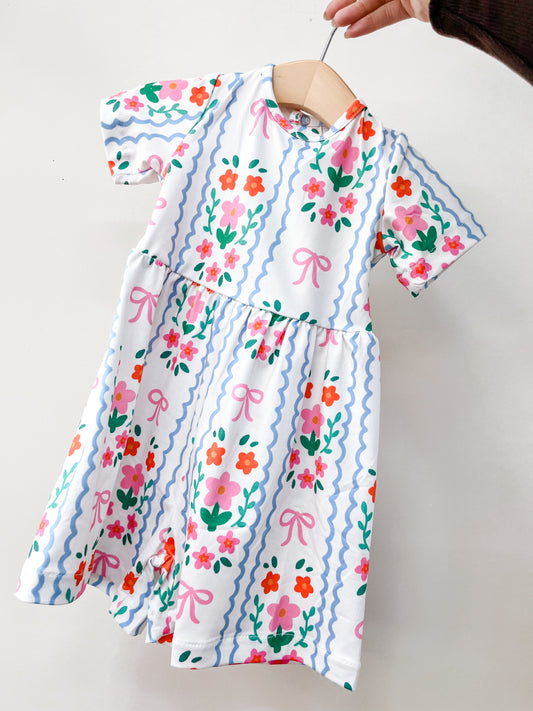 A hand holds up the Tiny Tot Bundles Ribbon Garden Romper, a short-sleeved white baby romper featuring colorful flowers, pink bows, blue wavy stripes, a gathered waist, and a vibrant design.