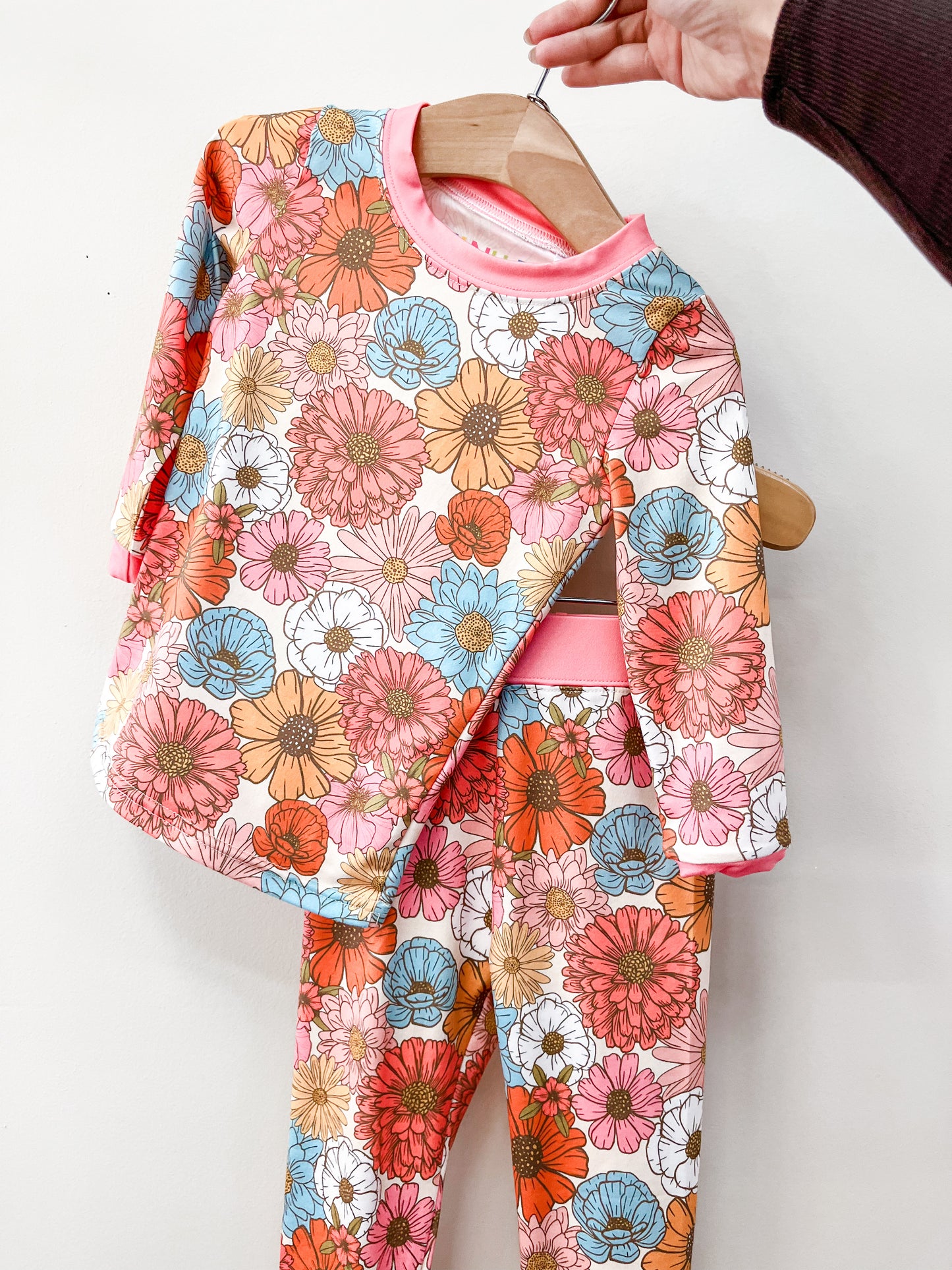A hand holds the Tiny Tot Bundles Retro Bloom Jammies Set—matching long-sleeve shirt and pants with a vibrant pink, orange, blue, and white floral pattern—displayed against a plain background.