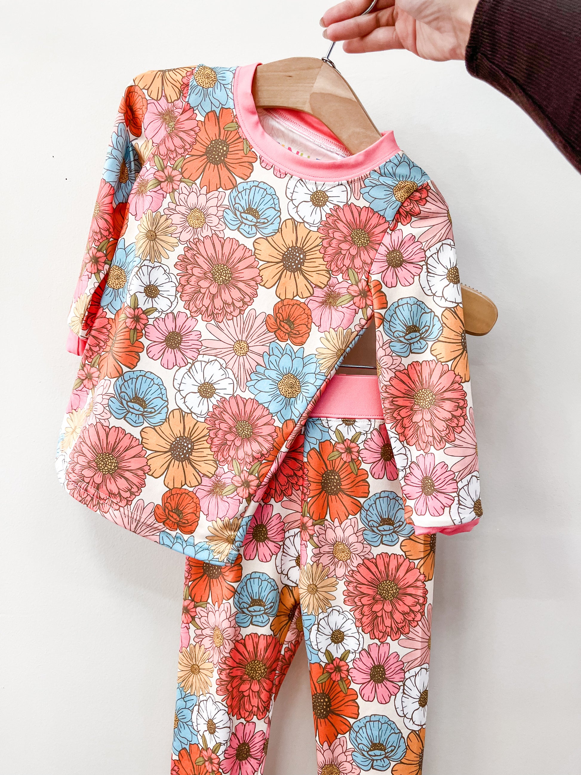 A hand holds the Tiny Tot Bundles Retro Bloom Jammies Set—matching long-sleeve shirt and pants with a vibrant pink, orange, blue, and white floral pattern—displayed against a plain background.