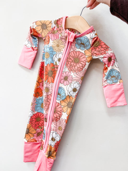 A hand holds the Tiny Tot Bundles Retro Bloom Zippy, a baby onesie with a colorful floral pattern in pink, orange, blue, and white plus pink cuffs and zipper, shown on a wooden hanger against a plain white background.