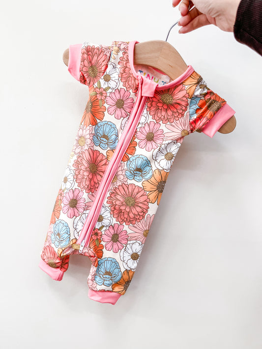 A hand holds the Tiny Tot Bundles Retro Bloom Shorty, a baby romper with a vibrant floral pattern in pink, orange, brown, and blue, displayed on a wooden hanger against a plain white background.