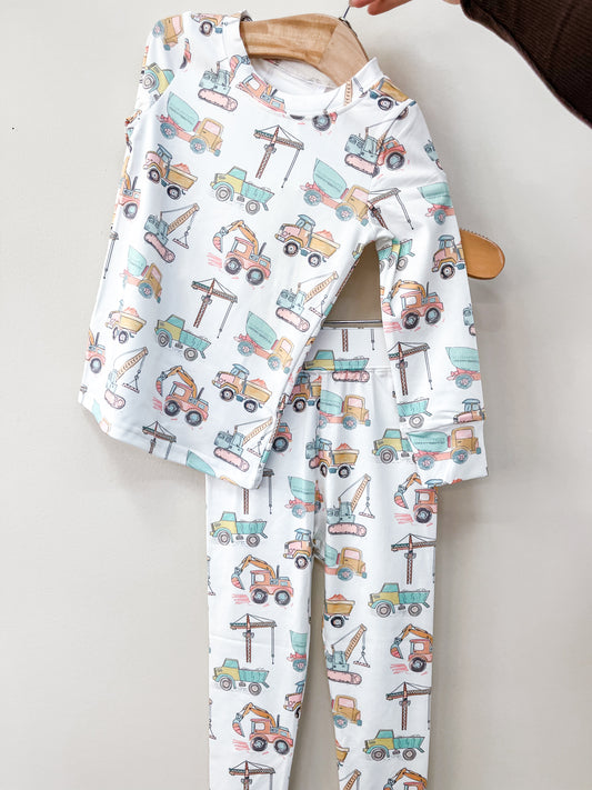 The Tiny Tot Bundles Construction Crew Jammies Set features long-sleeve pajamas and pants with colorful construction vehicles on a white background, displayed on a wooden hanger against a light wall.