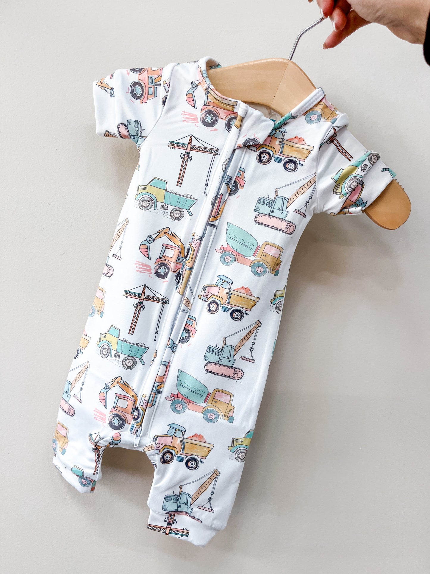 A hand holds a hanger displaying the Tiny Tot Bundles Construction Crew Shorty baby romper, featuring colorful prints of trucks, cranes, and diggers against a plain light background.
