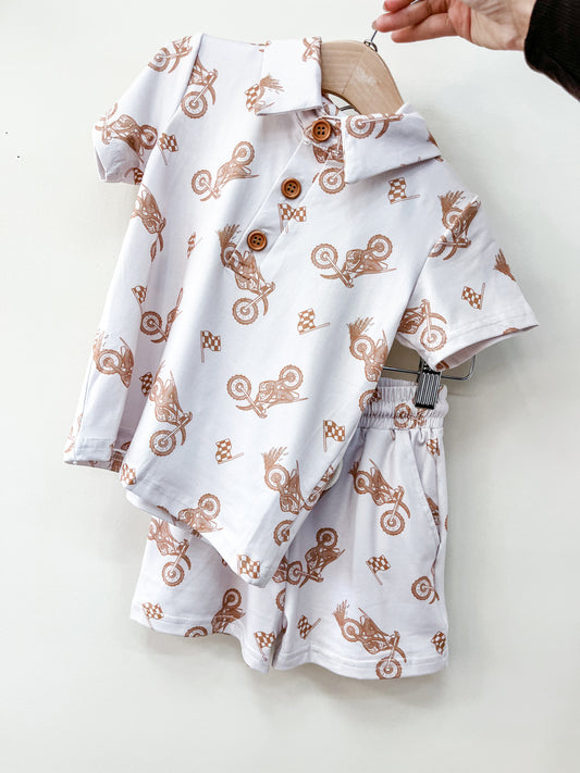 A hand holds the Tiny Tot Bundles Moto Ride Lounge Set: a white toddler outfit with a short-sleeve button-up shirt and matching shorts, both adorned with brown motorcycle and checkered flag designs.
