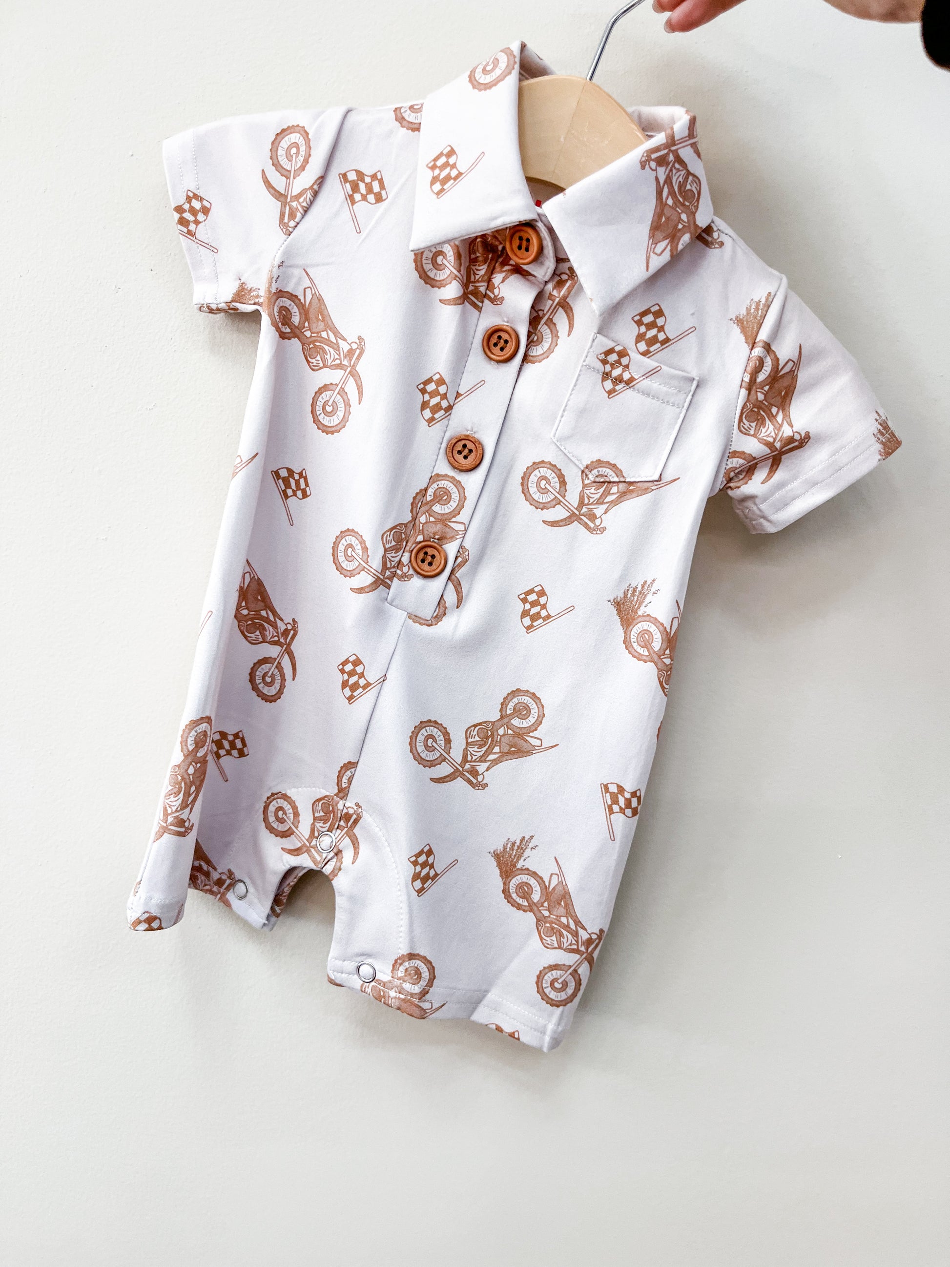 The Moto Ride Collard Romper by Tiny Tot Bundles is a short-sleeved white baby romper with brown motocross bike and checkered flag prints, wooden buttons, and a front pocket, displayed on a wooden hanger against a light background.