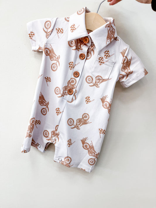 The Moto Ride Collard Romper by Tiny Tot Bundles is a short-sleeved white baby romper with brown motocross bike and checkered flag prints, wooden buttons, and a front pocket, displayed on a wooden hanger against a light background.