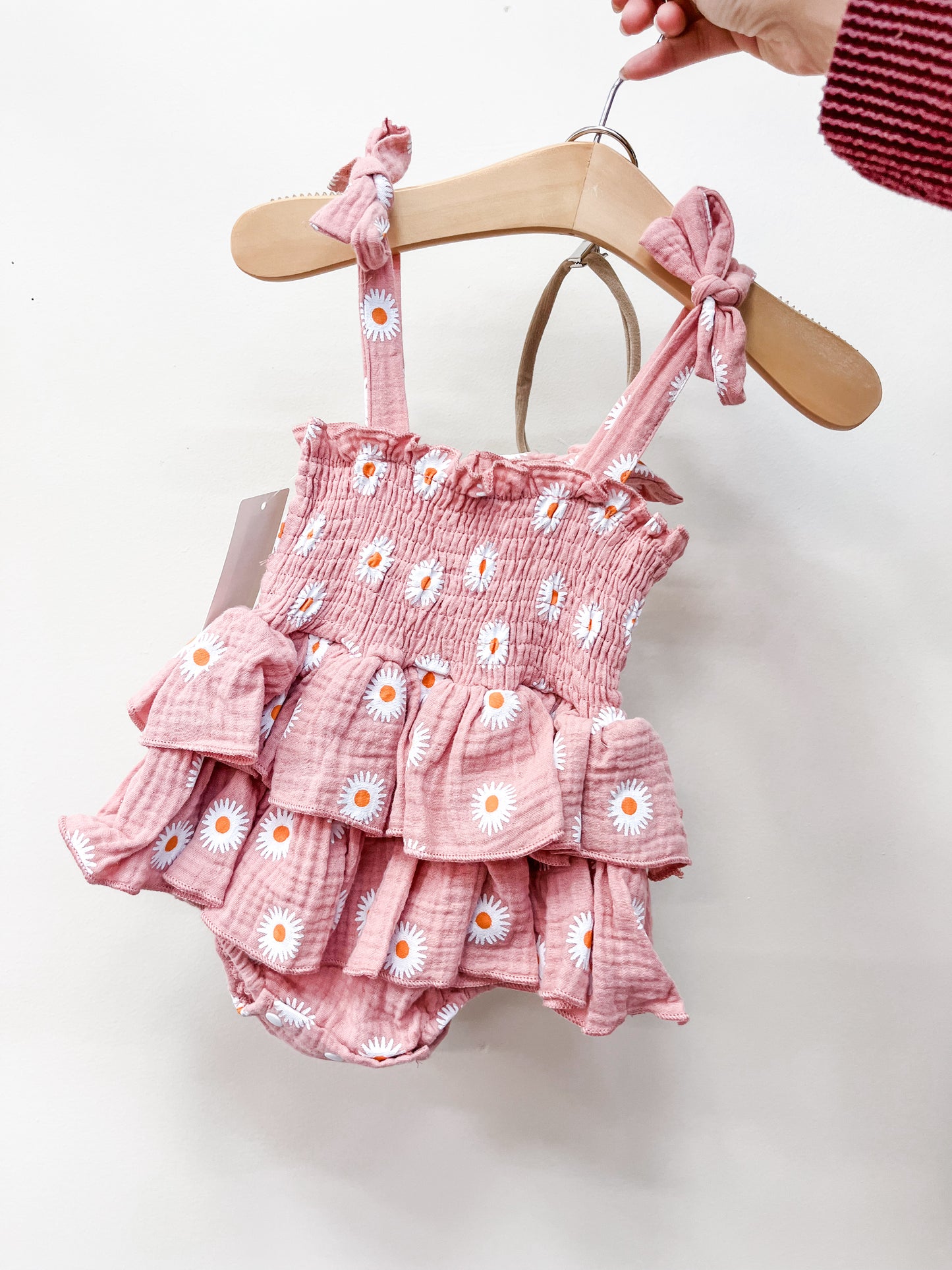 A hand holds a Tiny Tot Bundles Rosy Daisy Ruffle Bubble—a pink baby romper with white daisy prints, ruffled layers, and tied shoulder straps—displayed on a wooden hanger against a plain white background.
