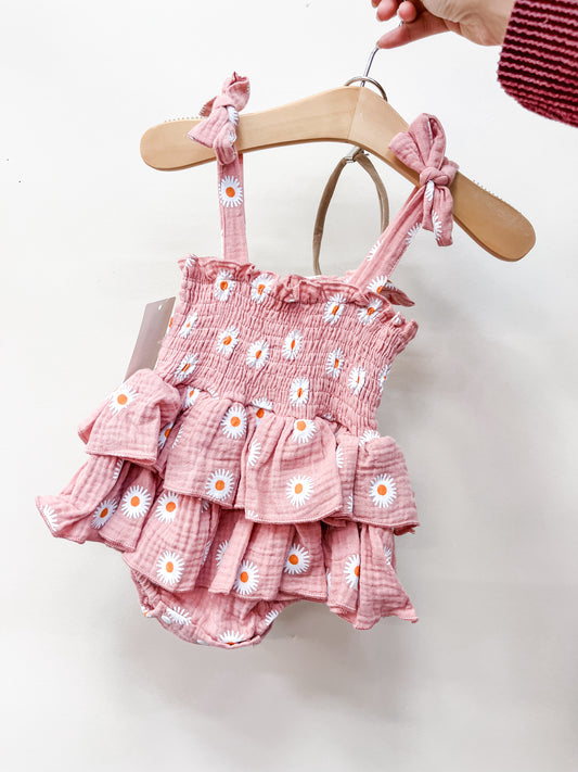 A hand holds a Tiny Tot Bundles Rosy Daisy Ruffle Bubble—a pink baby romper with white daisy prints, ruffled layers, and tied shoulder straps—displayed on a wooden hanger against a plain white background.