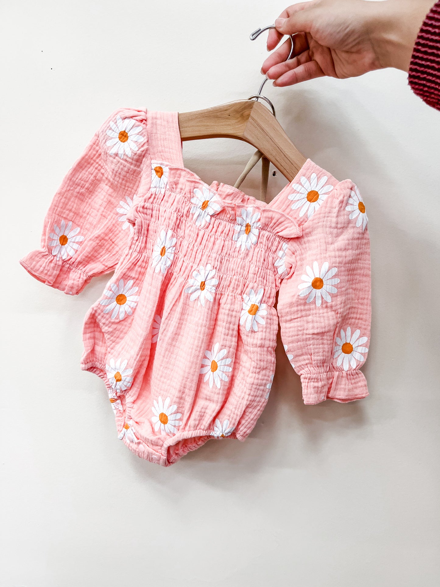 A hand holds the Tiny Tot Bundles Coral Daisy Romper, a pink baby outfit with white daisy prints and puffed sleeves, displayed on a wooden hanger against a plain light background.