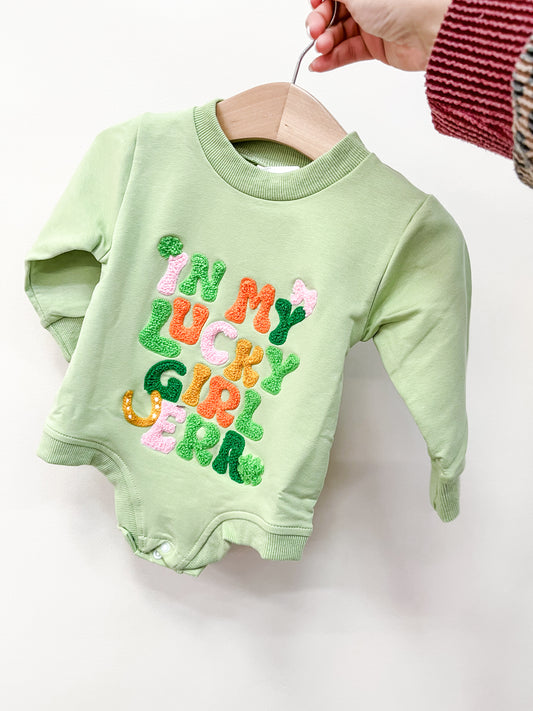 A hand holds the Green Lucky Girl Bubble by Tiny Tot Bundles on a wooden hanger. The front features colorful, textured "IN MY LUCKY GIRL ERA" lettering. The person wears a red and brown striped sweater.