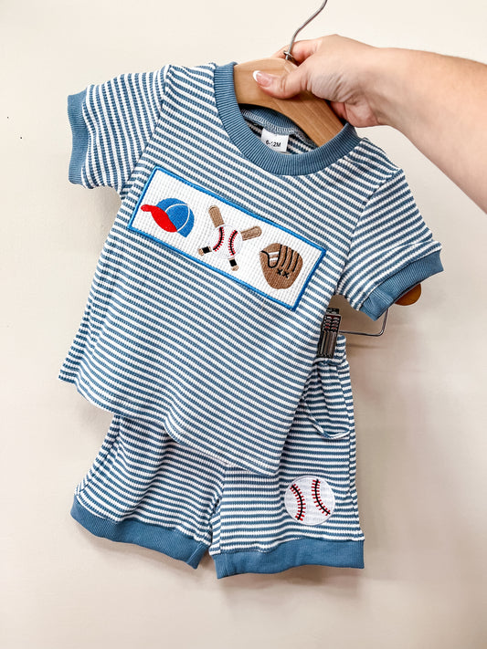 A hand holds up the Tiny Tot Bundles Blue Stripe Baseball Set, a toddler outfit with short sleeves and shorts featuring embroidered baseball gear on the shirt and two baseballs on the shorts.