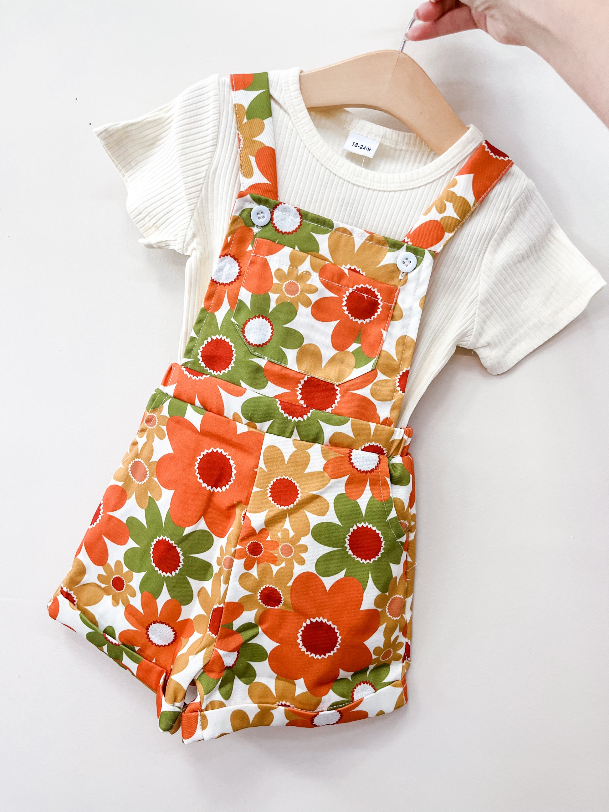 The Orange Blossom Overall Set by Tiny Tot Bundles, featuring a cream ribbed short-sleeve shirt and vibrant orange, green, and yellow floral overalls, is displayed on a hanger against a white background.