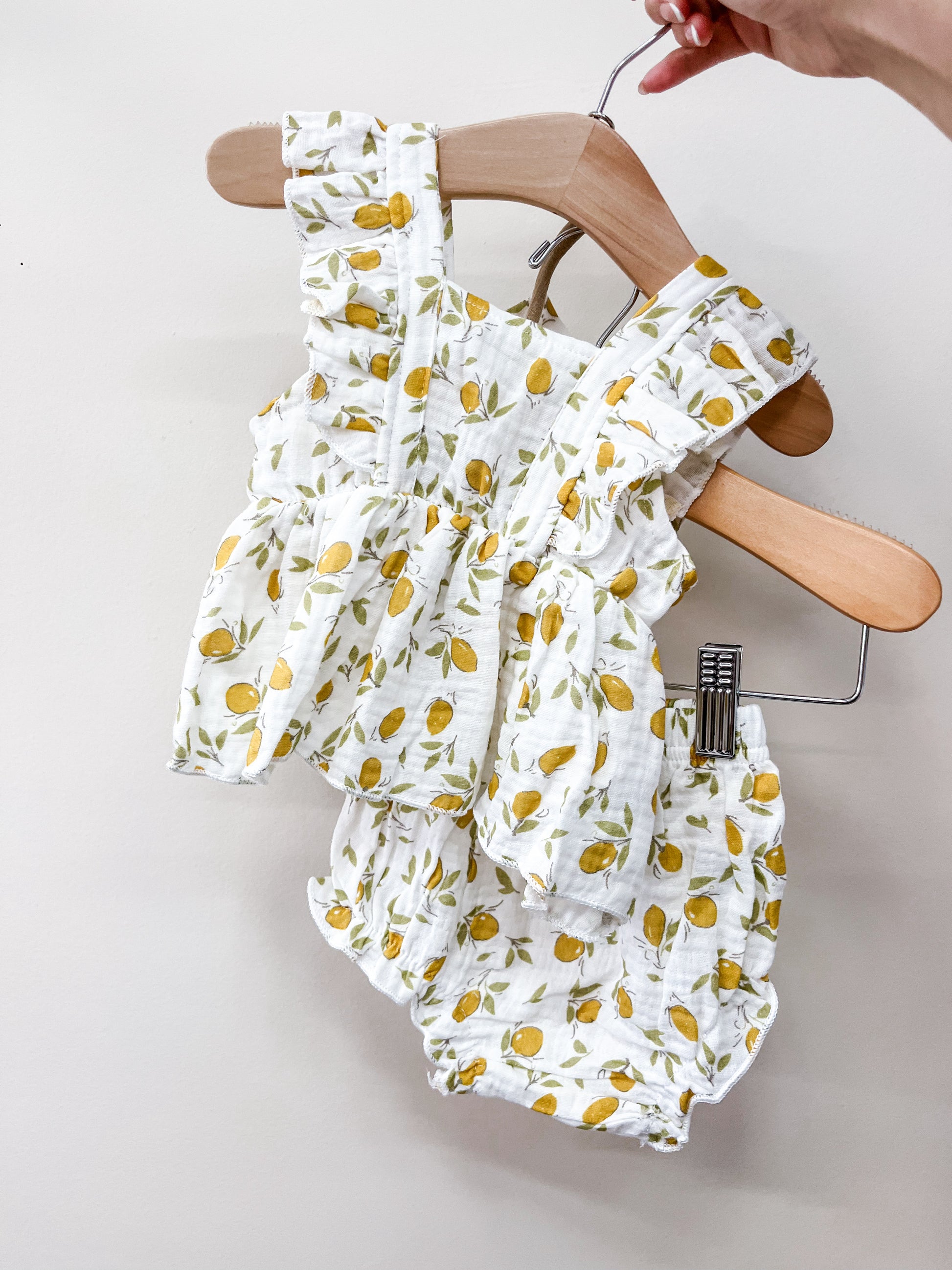 A hand holds the Tiny Tot Bundles Sunny Lemon Set, featuring a ruffled sleeveless top and matching bloomers with a yellow lemon print on a white background.