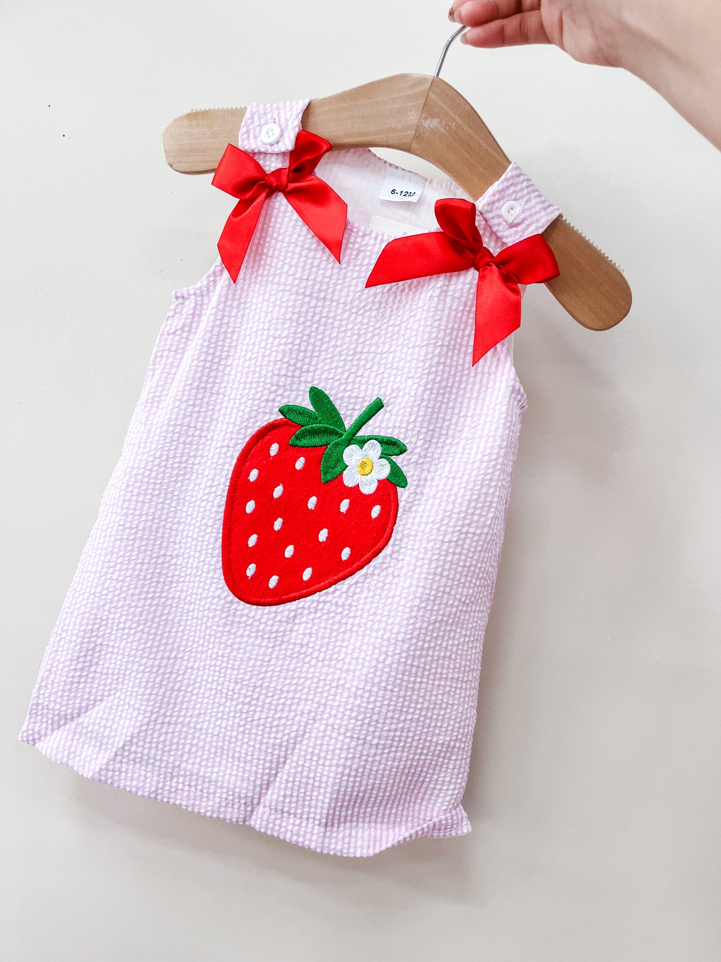 A hand holds a hanger with the Sweet Strawberry Dress by Tiny Tot Bundles, a sleeveless light pink textured dress adorned with a large strawberry applique on the front and two red bows on the shoulders.