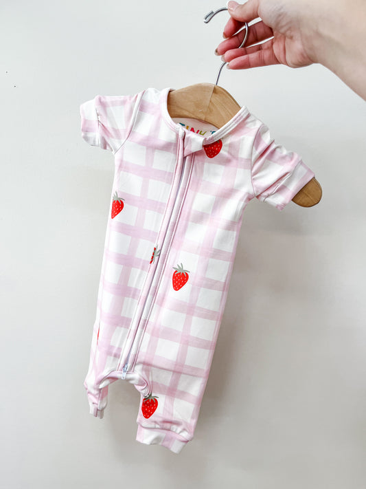 A hand holds a wooden hanger with the Tiny Tot Bundles Strawberry Picnic Zippy, a pink and white plaid short-sleeve baby romper with red strawberry prints and a front zipper, set against a plain light wall.