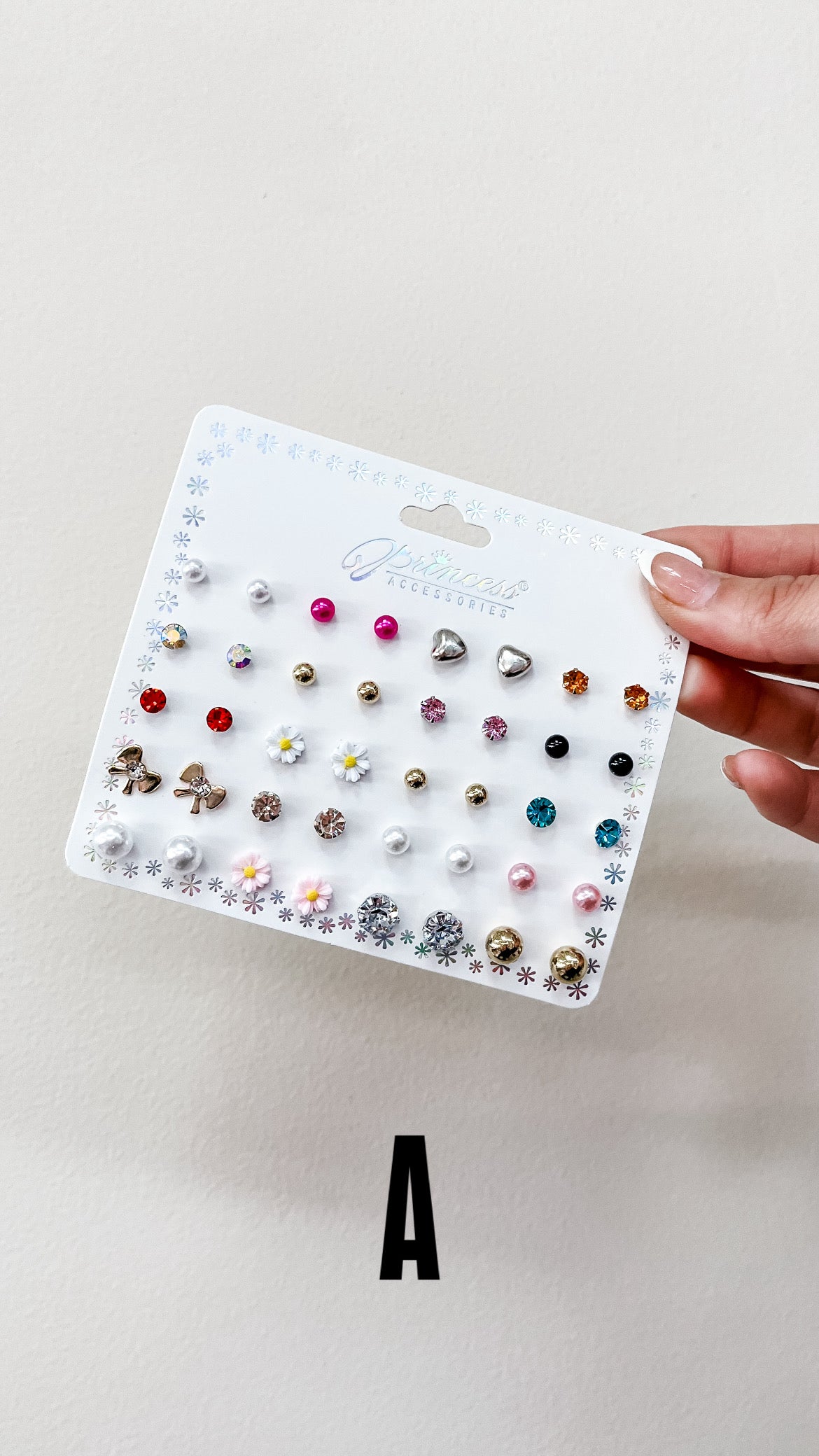 A hand holds a display card from Sensibling Corp.'s Mini Glam Stud Set, featuring colorful studs in shapes like flowers, stars, pearls, and rhinestones against a plain background. The letter A is printed below the card.