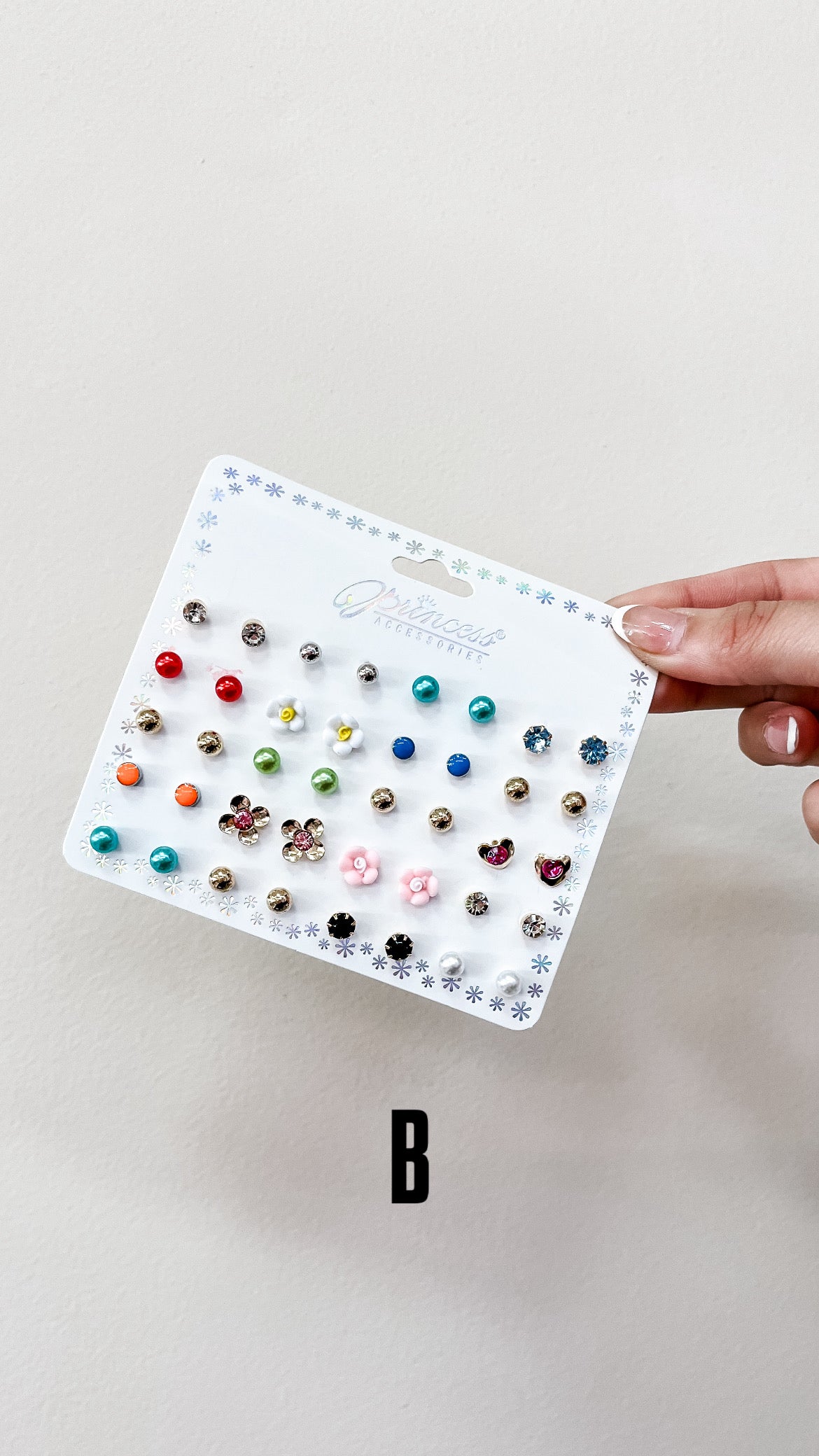A hand displays the Sensibling Corp. Mini Glam Stud Set—a white card with colorful flower, pearl, and gem stud earrings in various designs—against a plain off-white background, featuring a large "B" below.