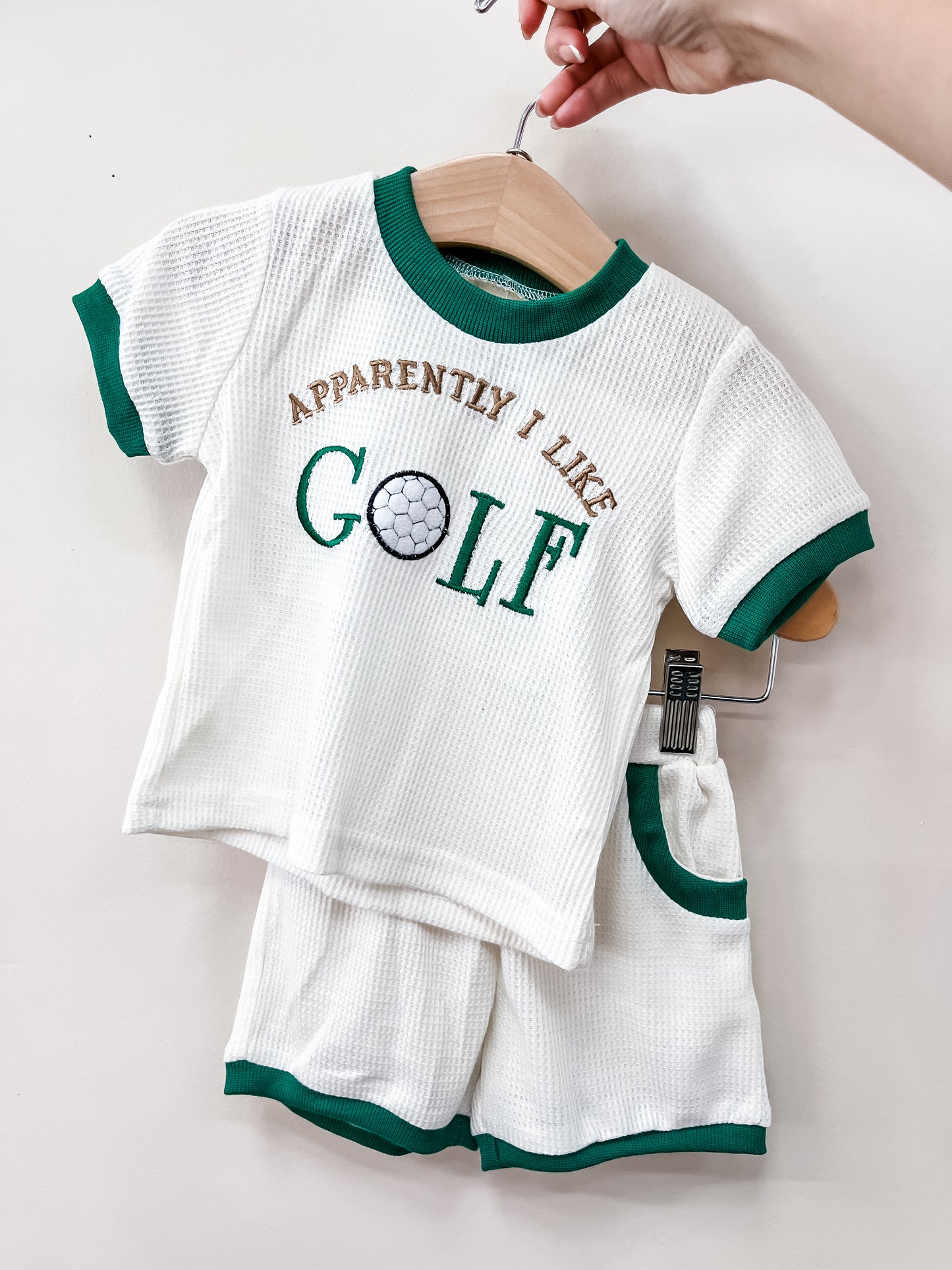 A hand displays the Tiny Tot Bundles "Future Golf Pro Set"—a white toddler outfit with green trim. The shirt reads "APPARENTLY I LIKE GOLF" with a golf ball design, and matching shorts hang below.