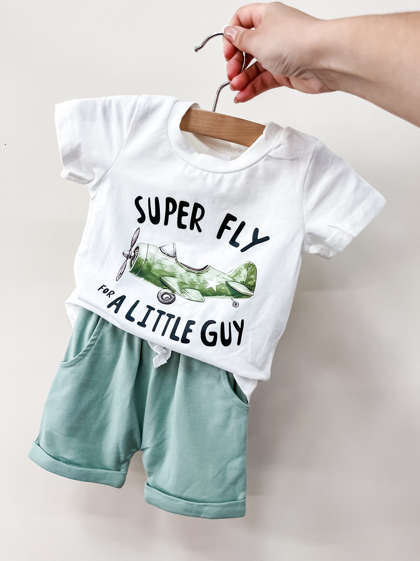 A hand holds up the Tiny Tot Bundles Super Fly Guy Set, featuring a white tee with a green airplane and "SUPER FLY FOR A LITTLE GUY" text, paired with mint green shorts.