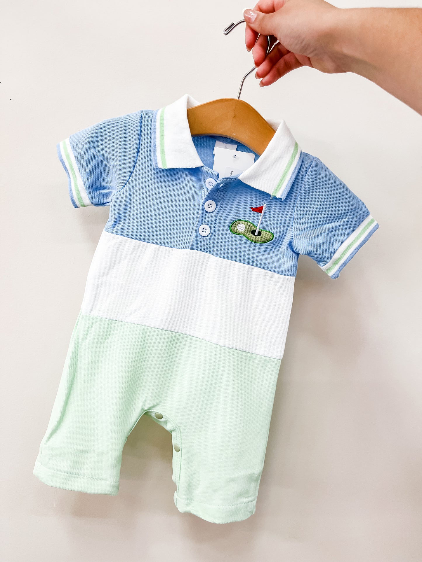 A hand holds a wooden hanger displaying the Tiny Tee Time Romper by Tiny Tot Bundles, a baby outfit with blue, white, and light green color blocks, a collar, and an embroidered golf green with a red flag on the chest.