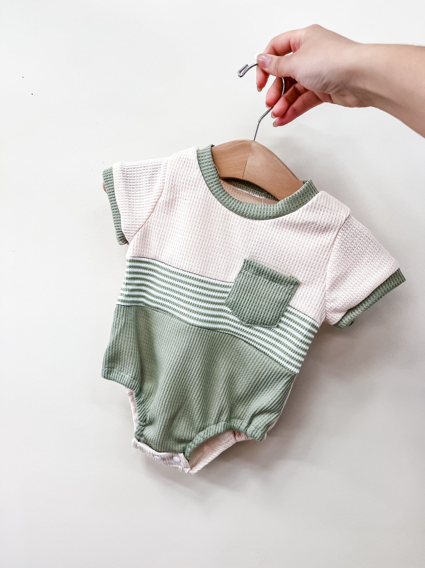 A hand holds a hanger with the Sage Coast Bubble by Tiny Tot Bundles, featuring a light pink top with green trim, a green pocket, green and white stripes across the middle, and a light green bottom.