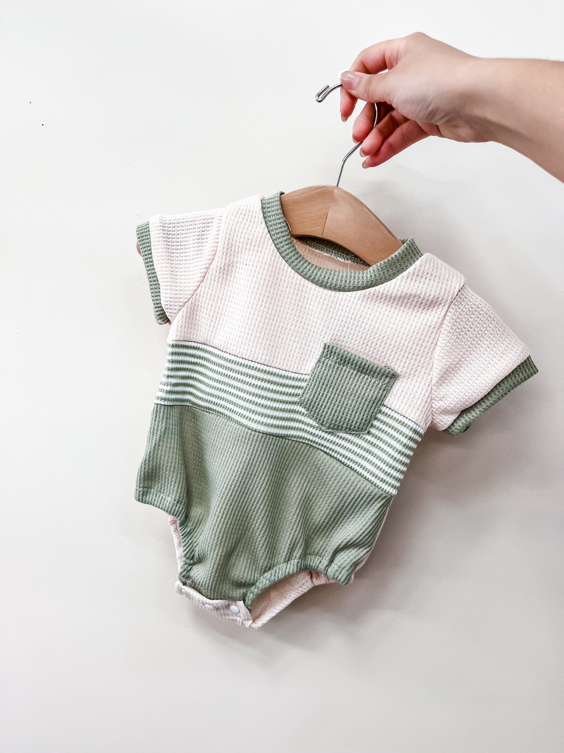 A hand holds a hanger with the Sage Coast Bubble by Tiny Tot Bundles, featuring a light pink top with green trim, a green pocket, green and white stripes across the middle, and a light green bottom.