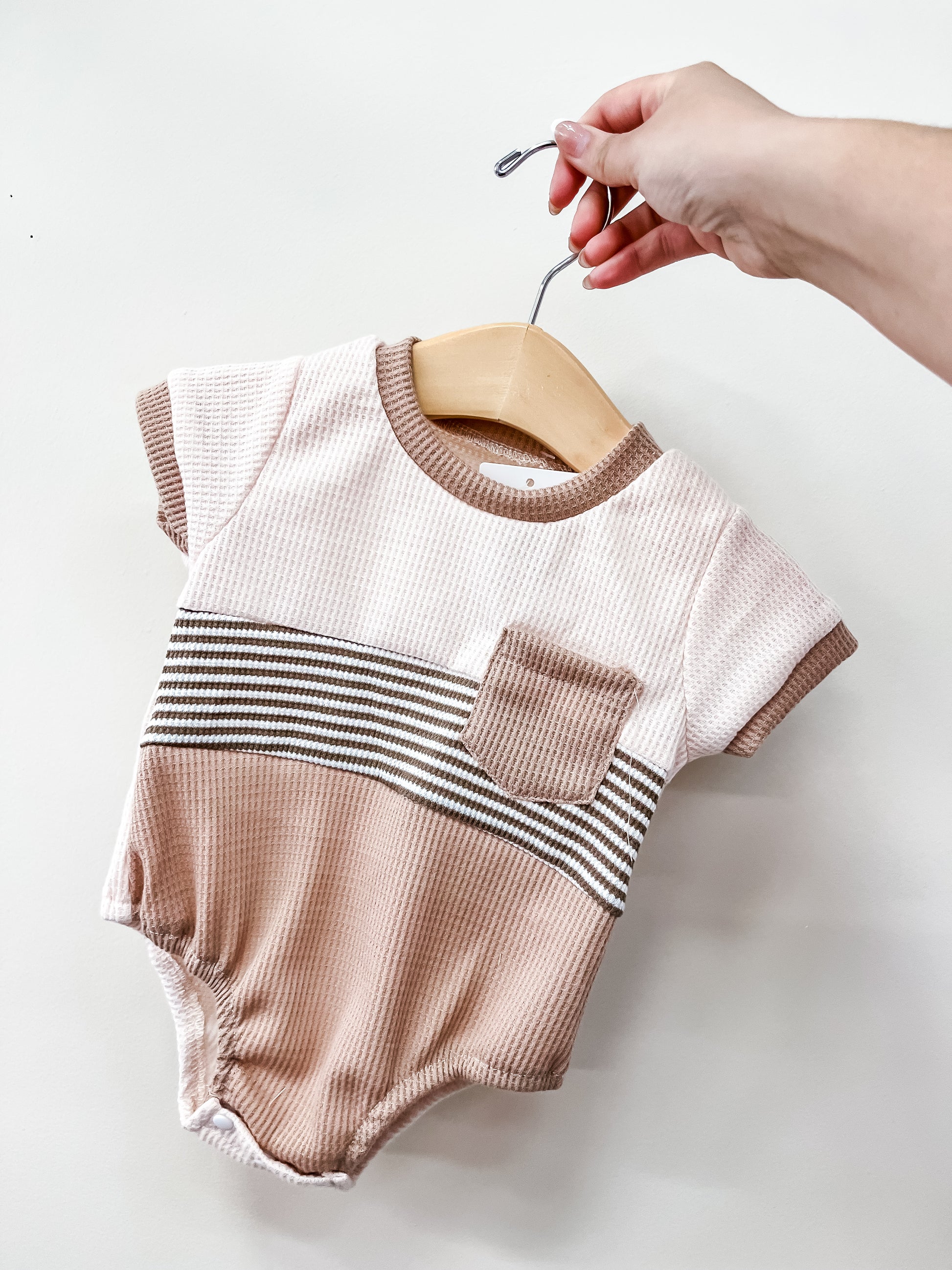 A hand holds the Cocoa Coast Bubble by Tiny Tot Bundles—a short-sleeve baby romper in beige and light brown stripes with a chest pocket—displayed on a wooden hanger against a plain white background.