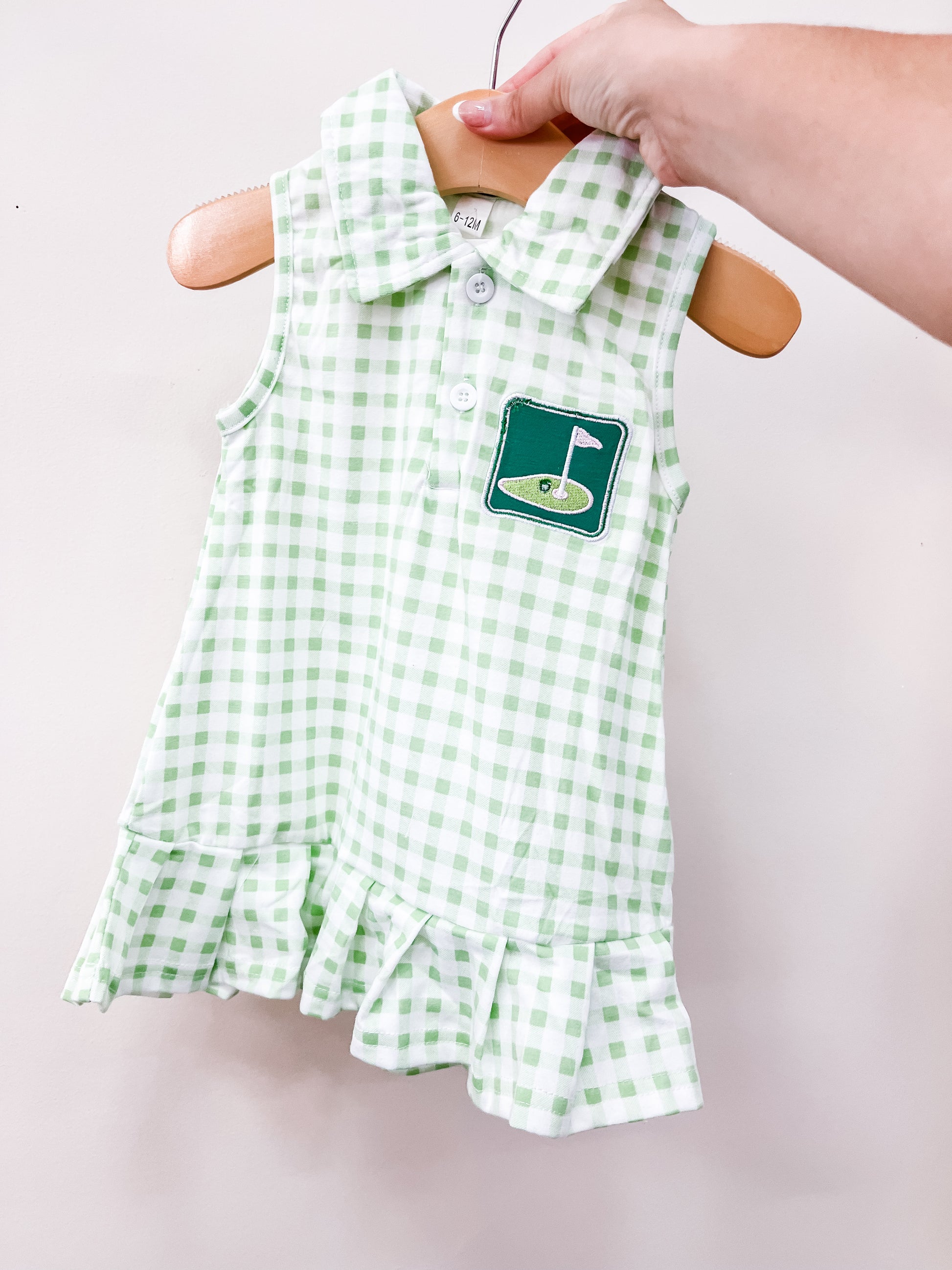 The Sweet Tee Gingham Dress by Tiny Tot Bundles is a sleeveless green and white gingham dress with a collar, buttons, golf flag chest patch, and ruffled hem, shown on a hanger held by hand.