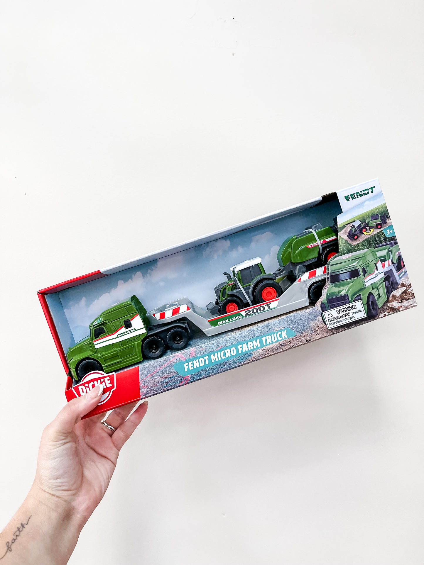 A hand holds a boxed Toy Smith Micro Farm Truck Assortment, featuring a green toy truck, trailer, and tractor inside clear packaging against a plain white background.