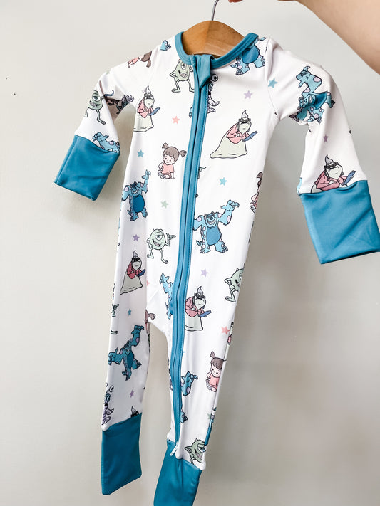 The Tiny Tot Bundles Monster Pals Long Zippy is a white baby onesie with teal cuffs and zipper, featuring cartoon Monsters, Inc. characters Sulley, Mike, and Boo, displayed on a wooden hanger against a white background.