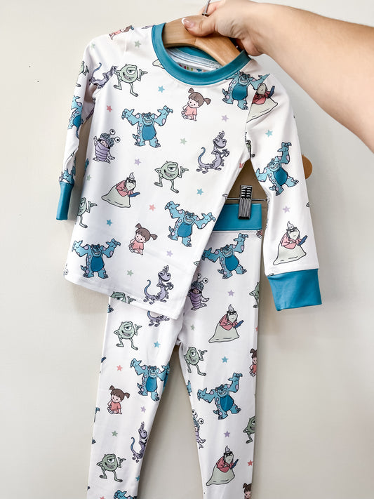 A hand holds up Tiny Tot Bundles' Monster Pals Jammies Set, a white pajama set with teal cuffs and an all-over print of Monsters, Inc. characters Sulley, Mike, Boo, and Randall.